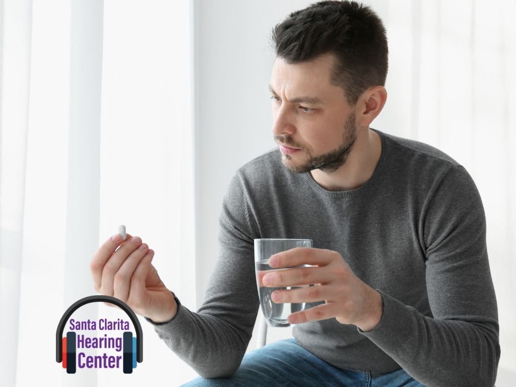 A person is holding a small white pill in one hand and a glass of water in the other. The person is wearing a gray long-sleeve shirt and blue jeans. The logo of 'Santa Clarita Hearing Center' is visible in the bottom left corner of the image, featuring a pair of headphones with the text 'Santa Clarita Hearing Center' inside. The image is likely related to health, medication, or hearing care.